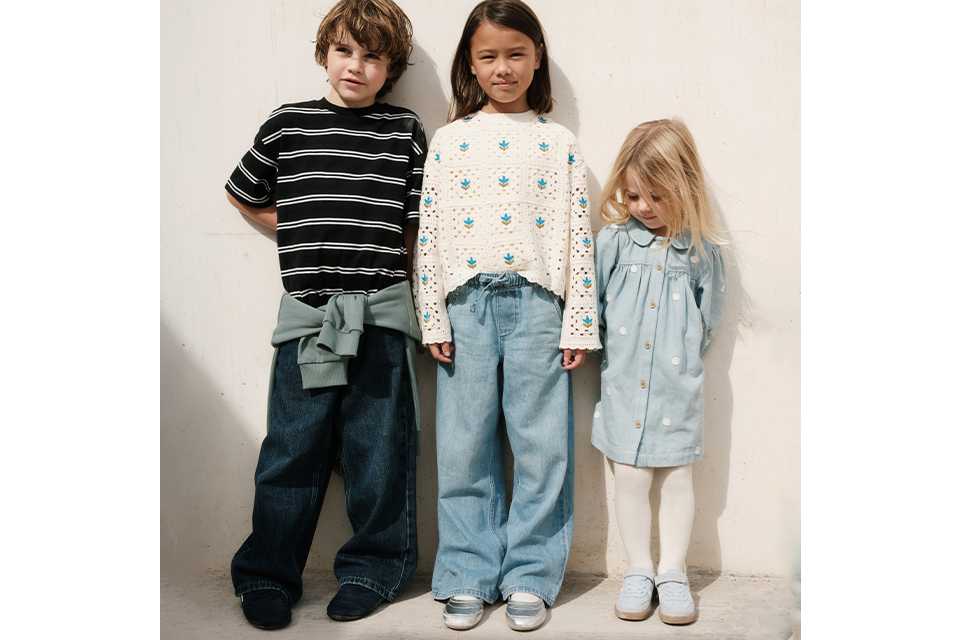 Three children standing against a light wall. One wears a black striped T-shirt with a green sweatshirt tied at the waist and dark wide-leg jeans. Another wears a cream sweater with a floral pattern and light blue wide-leg jeans. The third wears a light blue button-front dress with white tights and light blue shoes.