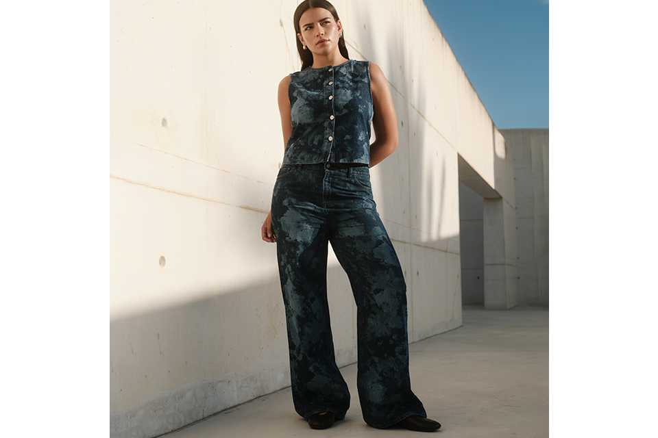 Person wearing a sleeveless button-front denim top with a dark blue tie-dye effect, paired with matching wide-leg denim jeans and black shoes, standing outdoors against a light concrete wall under a clear blue sky.