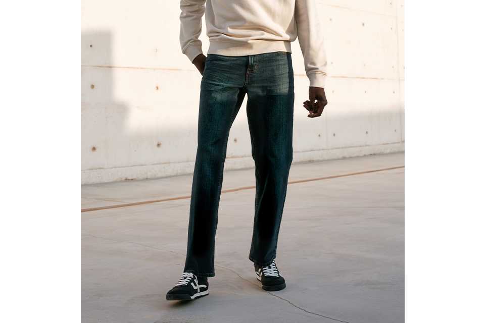 Person wearing a cream long-sleeve sweatshirt paired with dark blue straight-leg jeans and black-and-white sneakers, accessorized with a beige cap, standing outdoors against a light concrete wall under a clear blue sky.