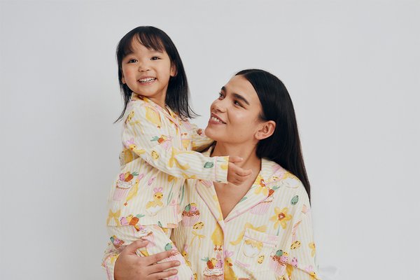 Two people wearing matching pink check pyjamas with small red heart patterns, sitting together on a light background.