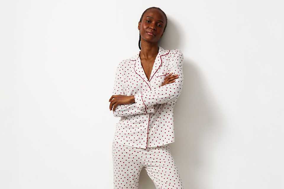 Person standing against a white wall wearing a white pajama set with small black and red heart prints, red piping detail, and beige fluffy slippers.