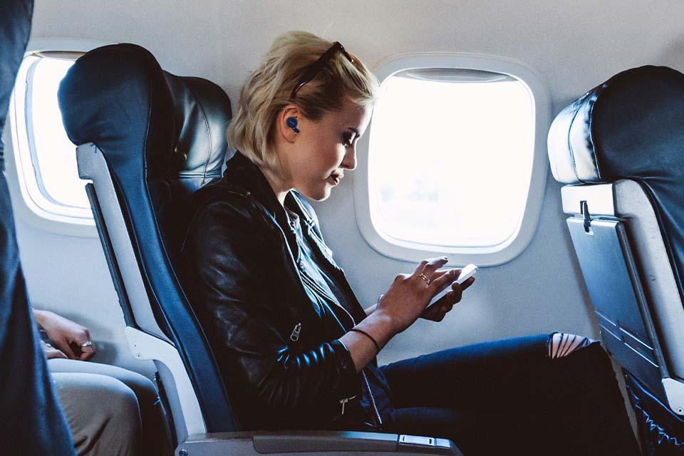 A lady sitting in an airplane seat listening to her phone through wireless earbuds.