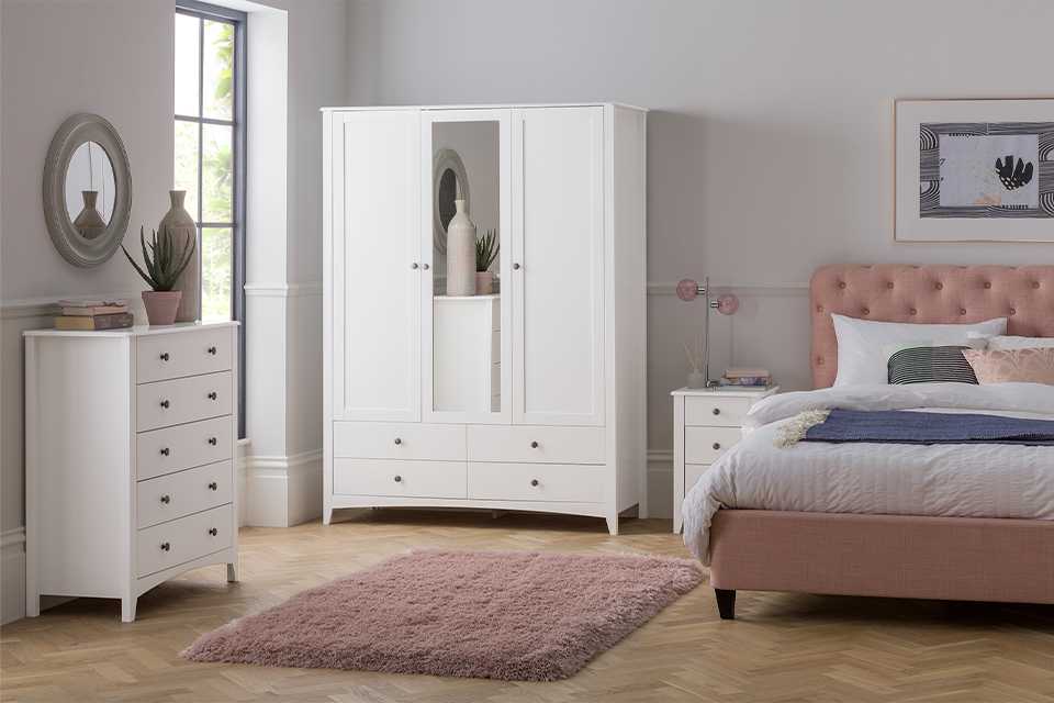 Image of white wardrobe, chest of drawers and two bedside drawers next to a pink bed.