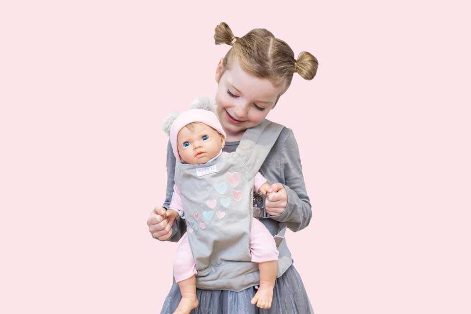 Image shows a small girl carrying a Tiny Treasures doll in a baby sling.