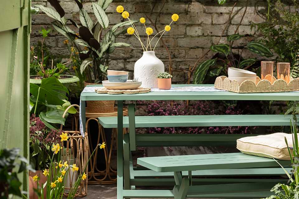 A green garden dining table set up with wicker accessories and an array of pots and planters.