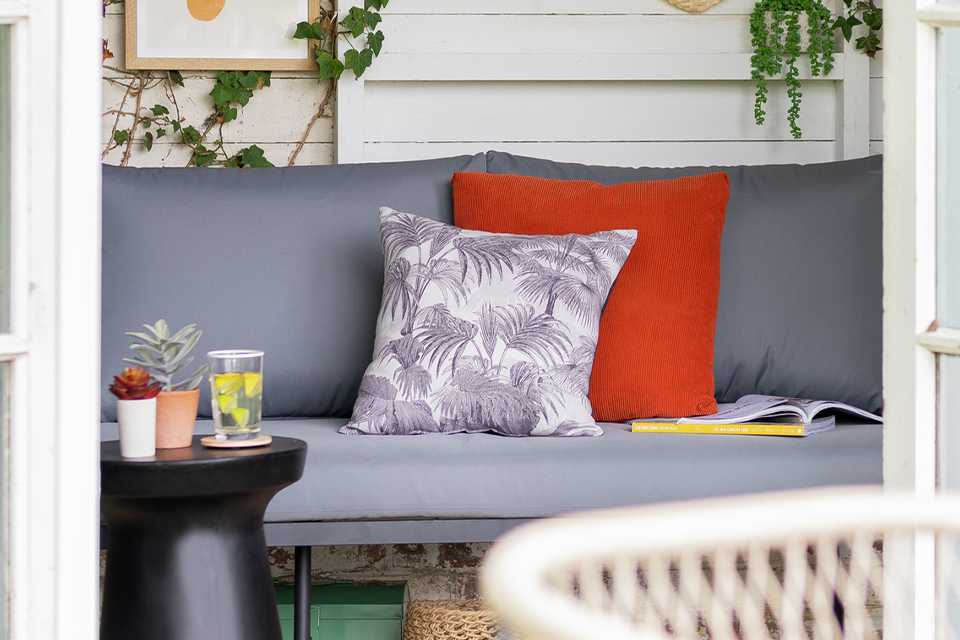 A grey outdoor sofa in a summerhouse with a grey leaf-print and a red cushion on it.