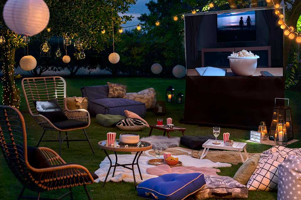 A garden set up for a movie night with a projector screen, cushions, blanket, garden chairs, beanbags, glassware, and serveware.