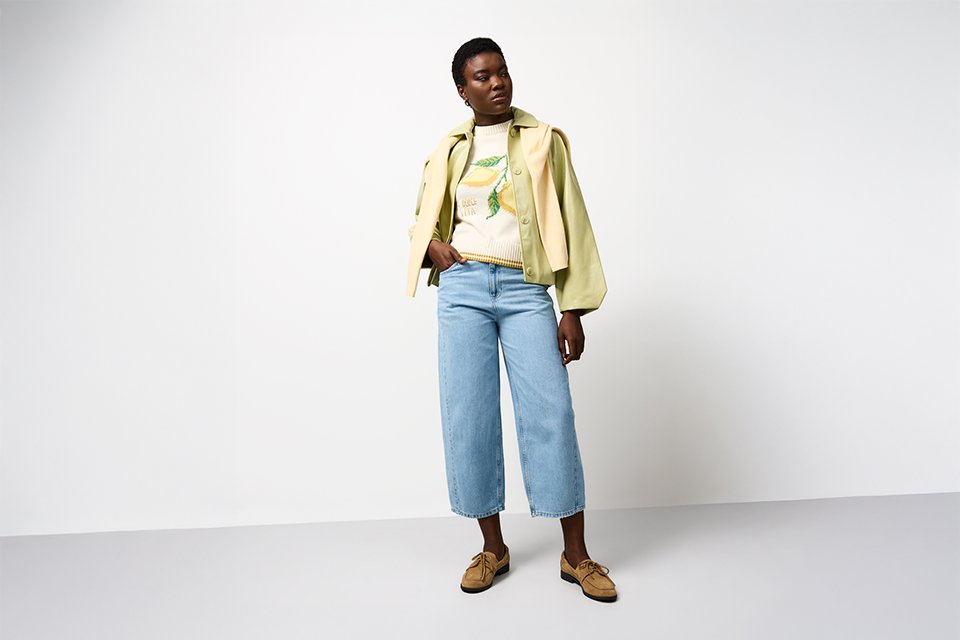 Model wearing a light green jacket layered over a printed top with cropped blue jeans and brown shoes, styled for spring layering.