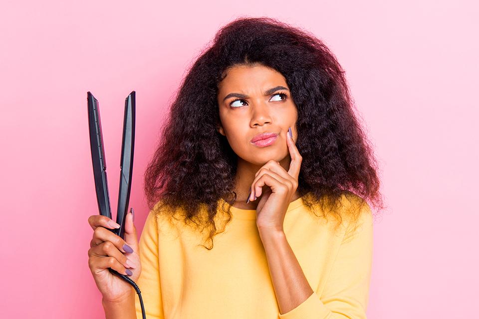 A lady holding up hair straighteners looking deep in thought.