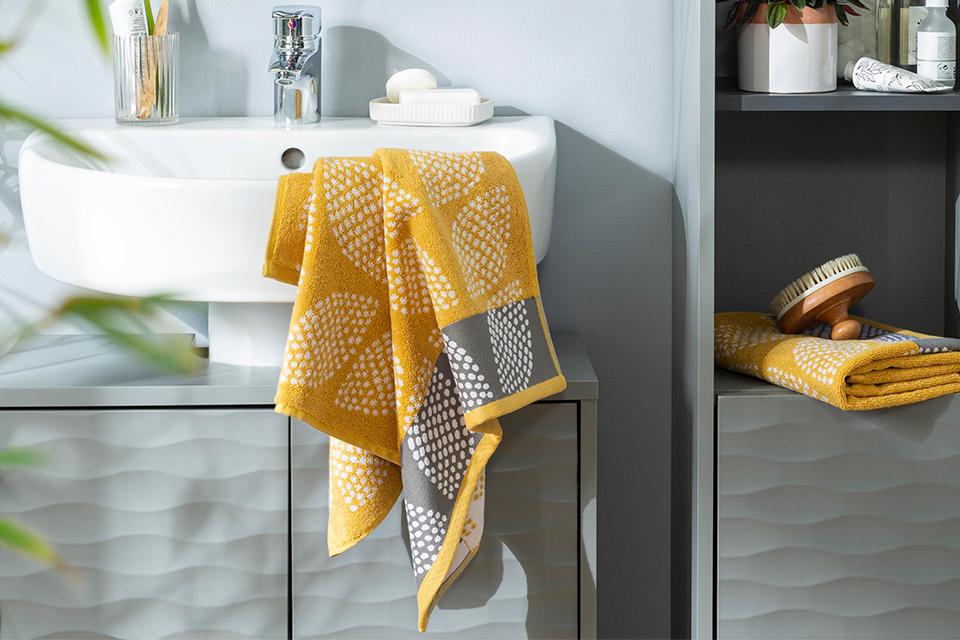 A yellow, grey and white leaf print towel displayed on a white sink and grey storage unit. 