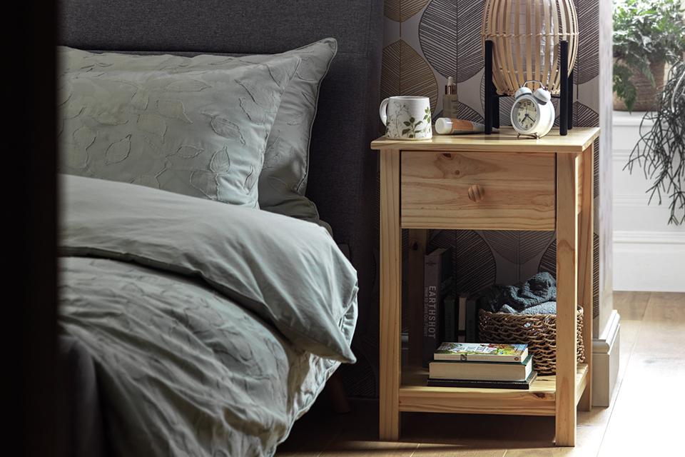 A wooden side table with one drawer next to a bed.