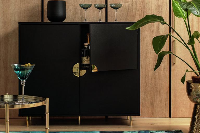 A Habitat 3 door drinks cabinet in black and gold finish with a vase and wine glasses on it and a drinks trolley in front of it. 