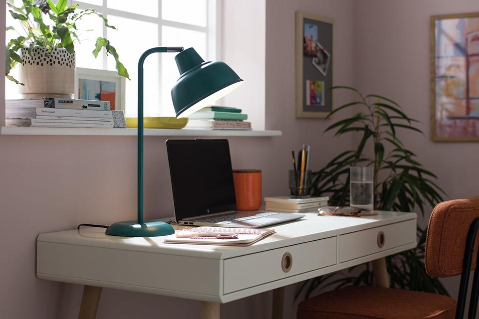 A bue desk lamp on a white and natural finish study table in a pink bedroom.