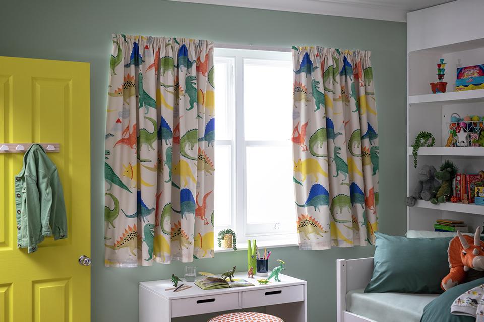A kids bedroom with white furniture and multi-coloured dino print curtains.