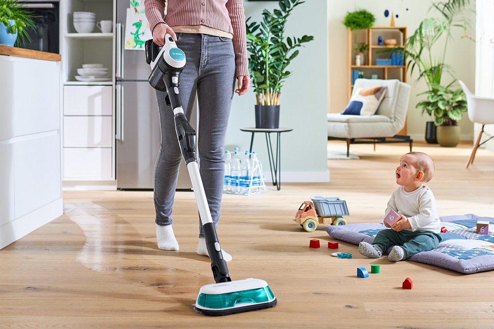A mum using a Bosch cordless vacuum cleaner to clean the floor with her baby on a playmat beside. 
