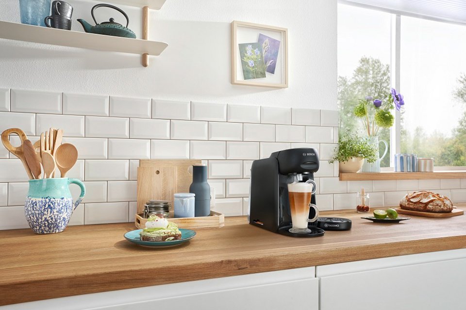 A black Tassimo by Bosch pod coffee machine with food items and crockeries on a kitchen countertop. 