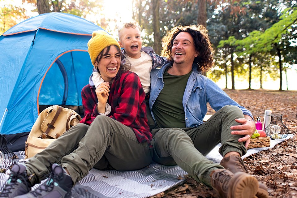 A family of 3 smiling and enjoying outside their tent.