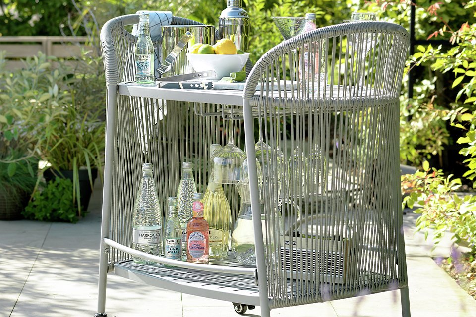 Grey wicker outdoor bar cart with bottles in garden.