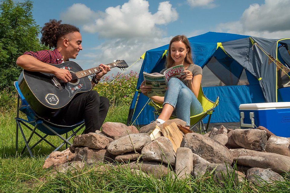 two people sit by a camp fire with a tent in the background