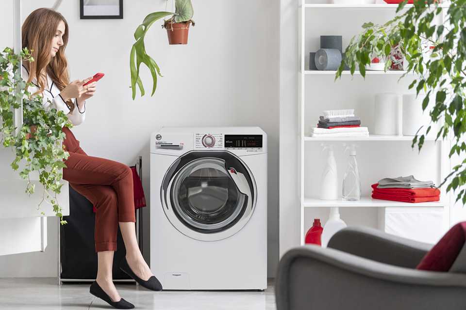 A lady on her mobile sitting by a washing machine waiting for it to finish.