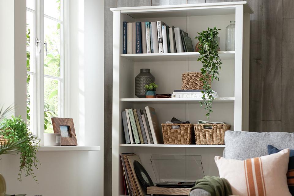 A white book shelf in a living room with books, storage baskets, vase and indoor plants on it.