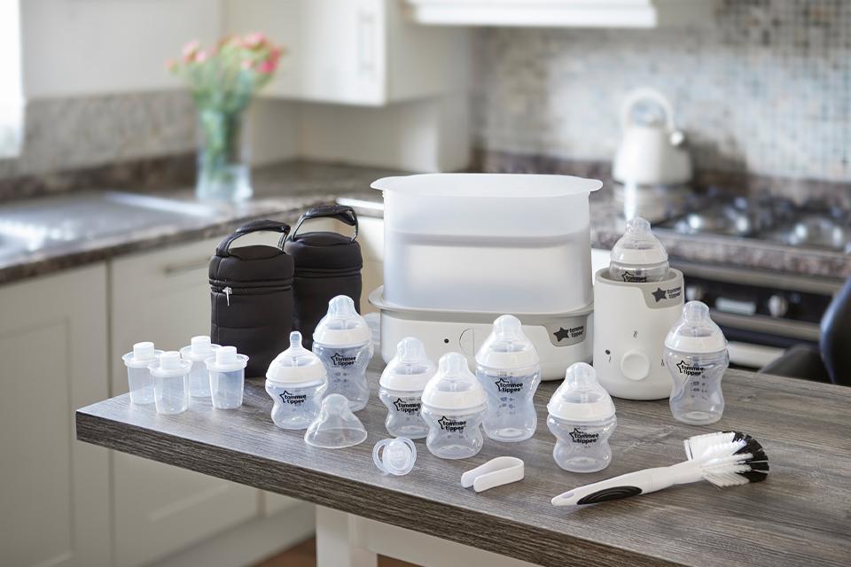 Image shows an assortment of Tommee Tippee bottles, sterilisers, cleaning brushes and accessories, laid out on a kitchen worktop. 
