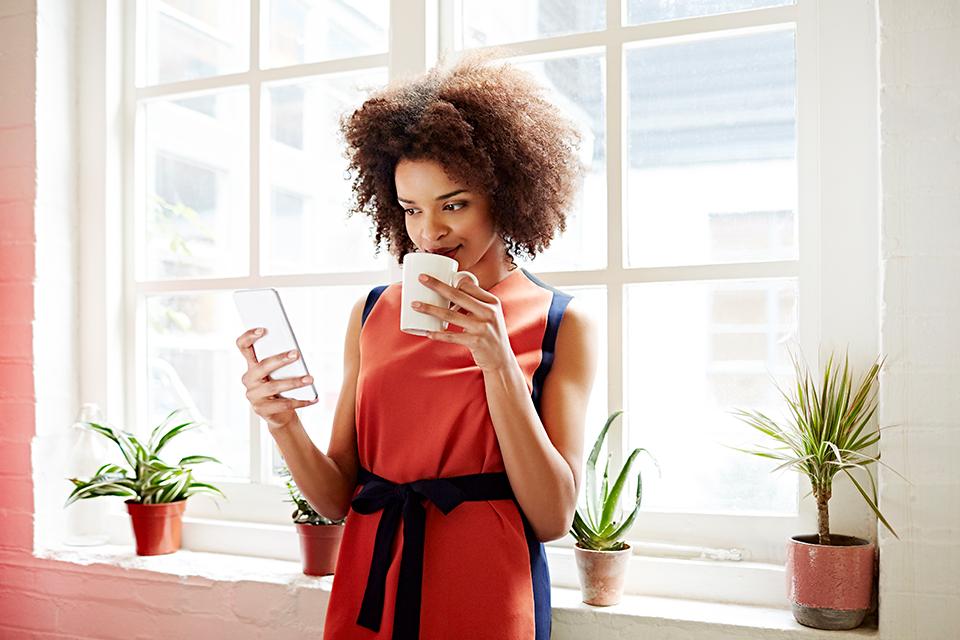 A woman drinking a cup of tea looks at her phone.