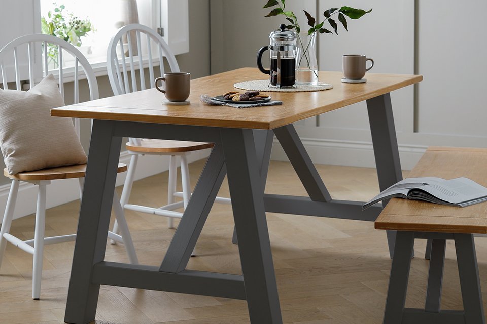 A grey Habitat solid wood 4 seater dining table with some coffee and a plant next to a window.