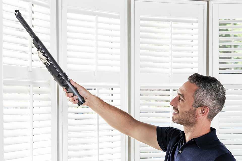 Man using a cordless, handheld vacuum cleaner to clean blinds.