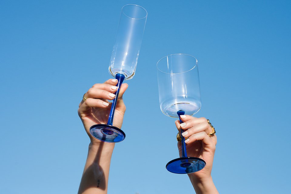 Hands holding two Habitat blue stemmed glasses above some grass outdoors.