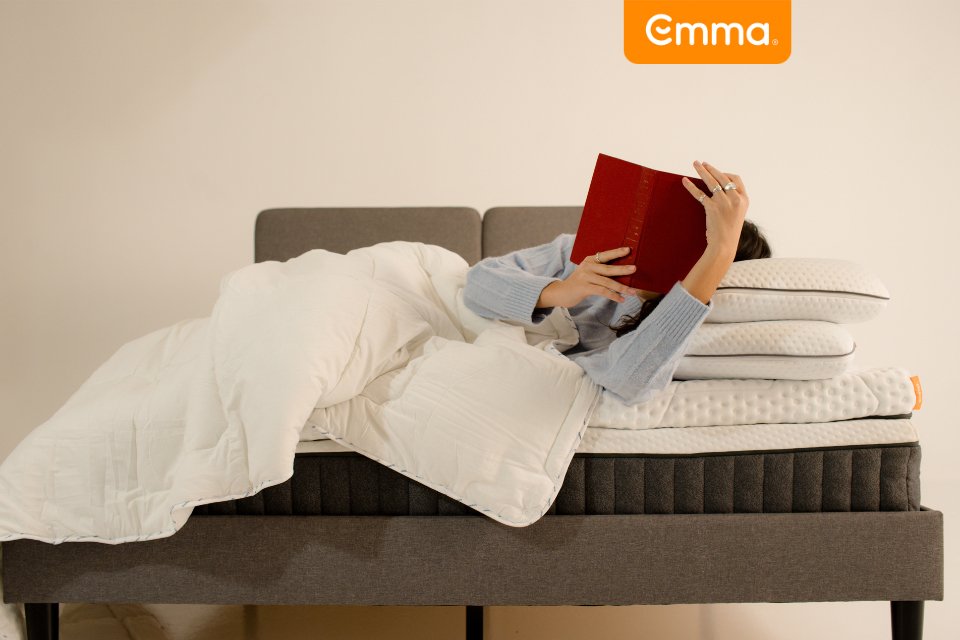 Person lying on an Emma mattress with pillows and duvet, reading a book on a bed.