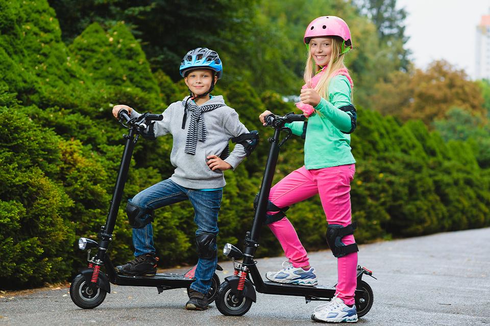 A boy and girl on electric scooters with a helmet, knee and elbow pads on.
