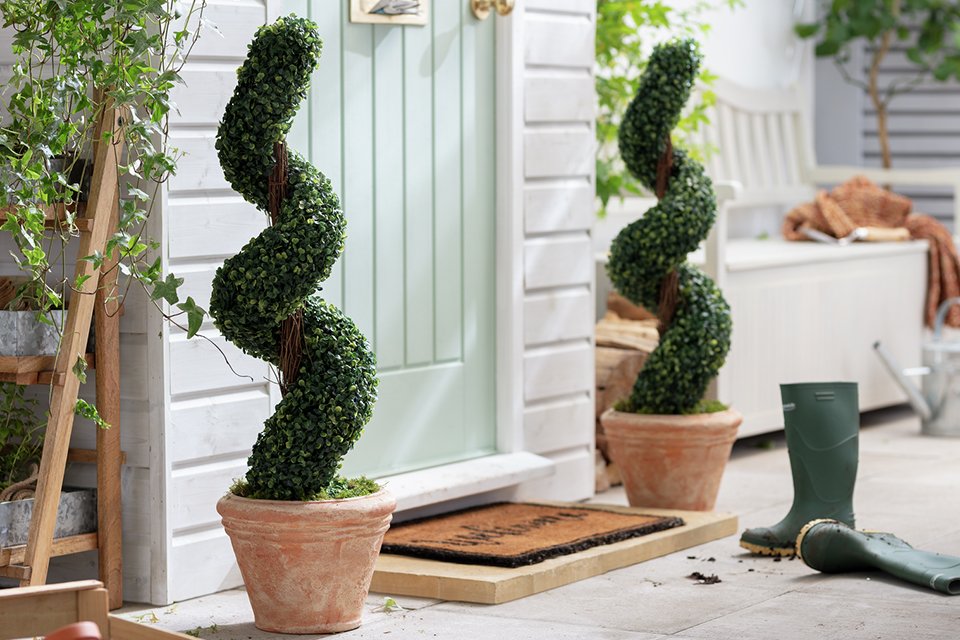 Welcome door mat in front of front door with faux spiral plants in terracotta plant pots either side of door.
