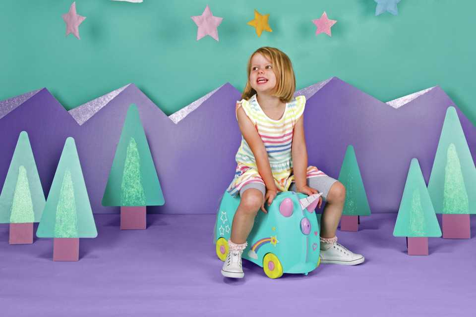 A girl sitting on a Trunki Una unicorn 4 wheel hard ride on suitcase in turquoise colour.