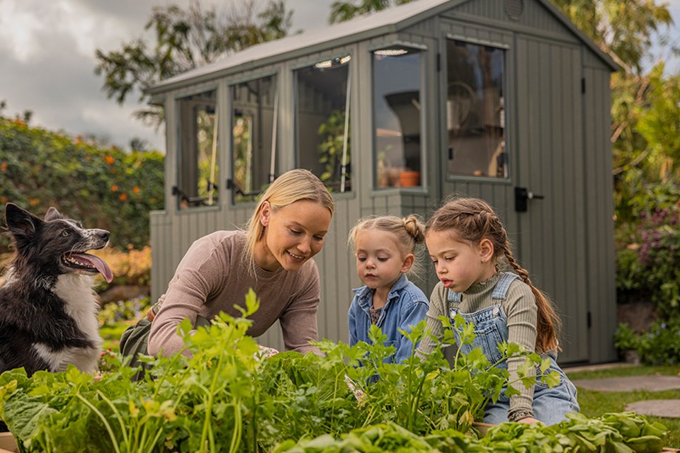 A woman, dog, and two children are planting shrubs in front of Keter Darwin Potting Shed.