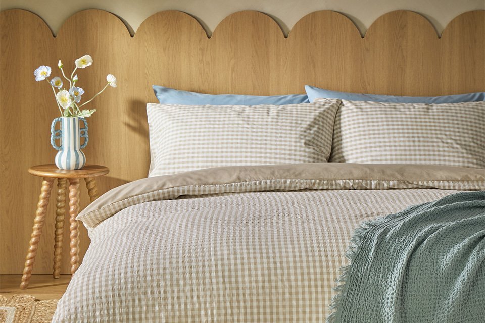 A Habitat gingham seersucker neutral bedding set on a bed next to a bedside table with flowers.