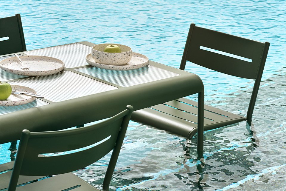 Three Habitat green outdoor chairs around a table with a white dinner set on it placed in a pool.