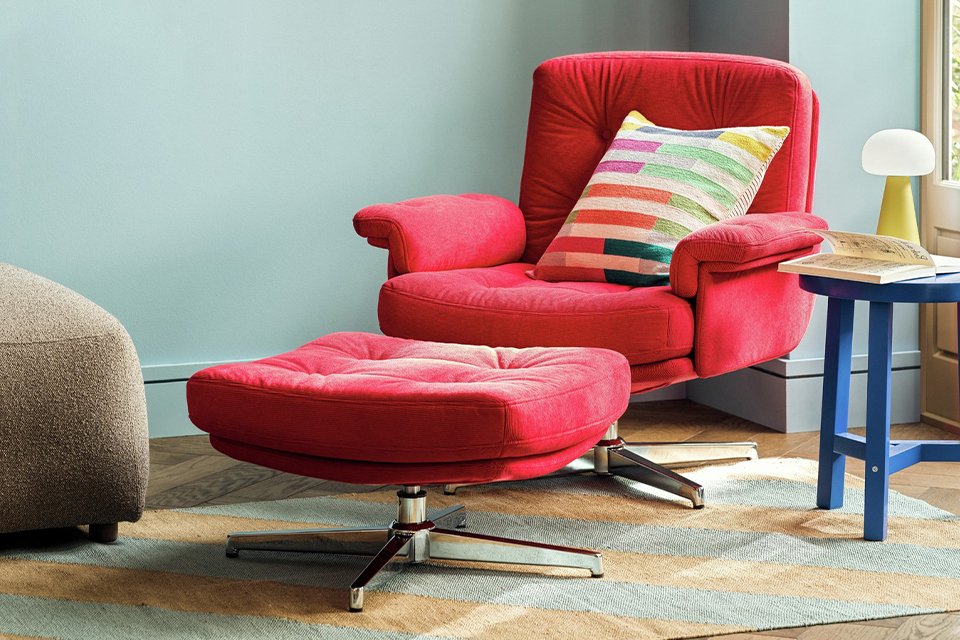 A red Habitat Dawson Fabric swivel chair with a colourful cushion next to a table with a lamp in a living room. 