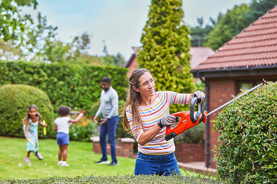 A woman using a Flymo® 18V EasiCut hedge trimmer to cut a garden hedge. 