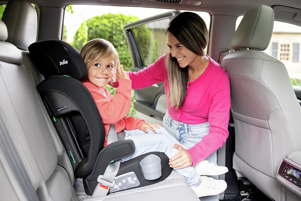 A mother strapping her child in a Joie Elevate R129 Toddler to Booster Car Seat in a car.