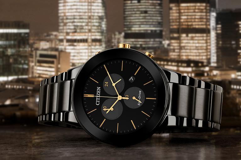 Image shows a sleek black Citizen watch with gold accents, against a background of glittering skyscrapers at night.