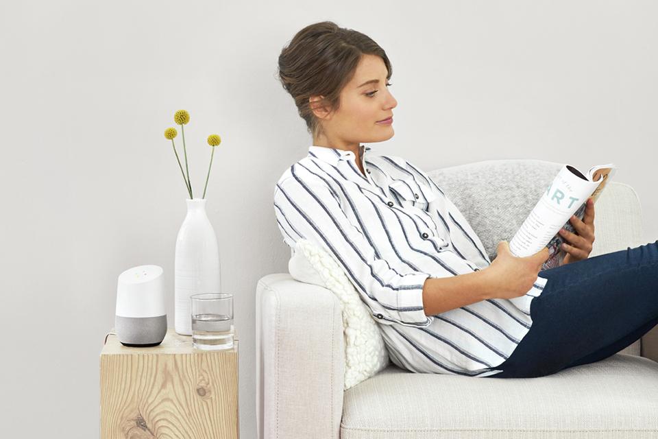 A smart speaker is placed next to an arm chair where a lady is sat reading.