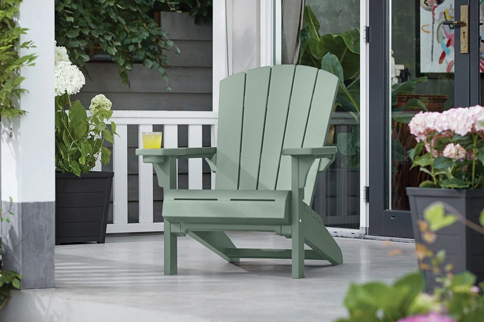 A green Keter Alpine Adirondack resin chair placed outdoors near green plants and flowers.