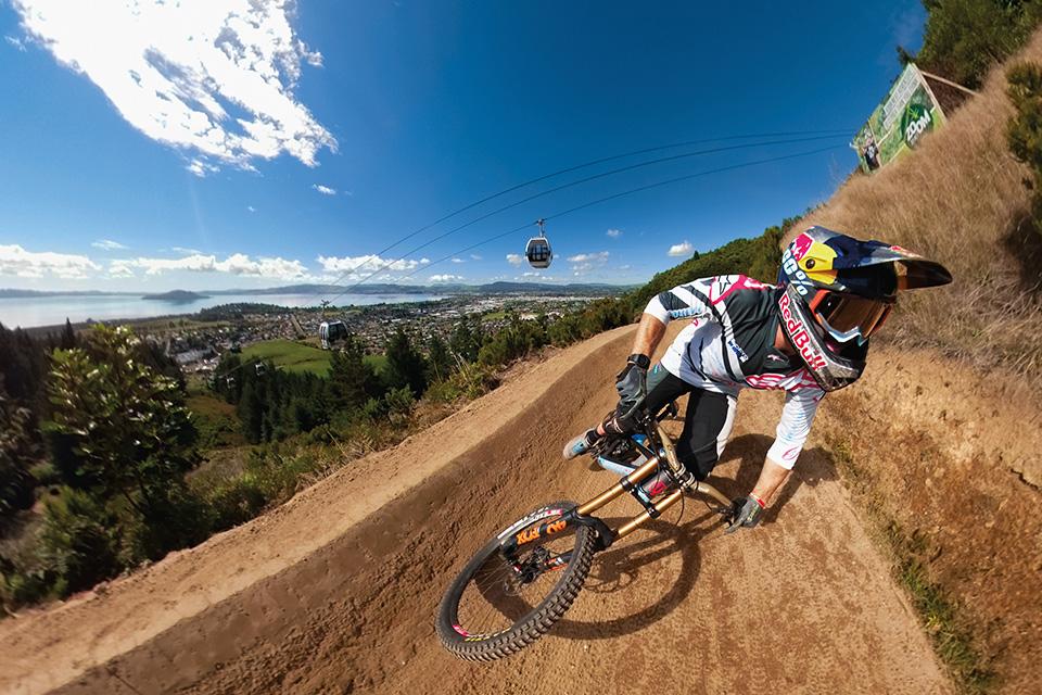 Image shows a helmet-clad cyclist riding a dirt track at a steep incline. Behind him, the landscape stretches out to.