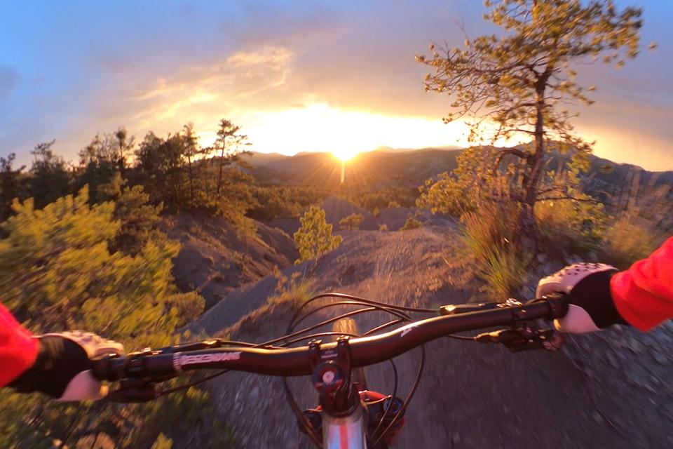 A rocky mountain landscape is seen from the point of view of a mountain biker, whose hands and handlebars point towards a flare of golden light.