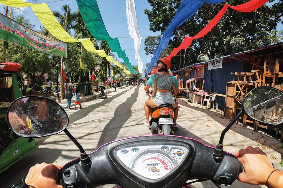 A sunny South East Asian street scene is viewed from behind the handlebars of a small motorbike. The wide-angle view creates a panoramic effect.