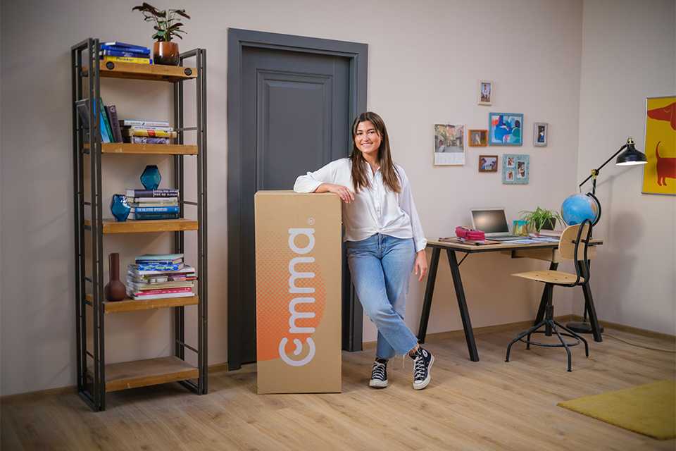 A woman standing next to an Emma Sleep cardboard box containing an Emma mattress.
