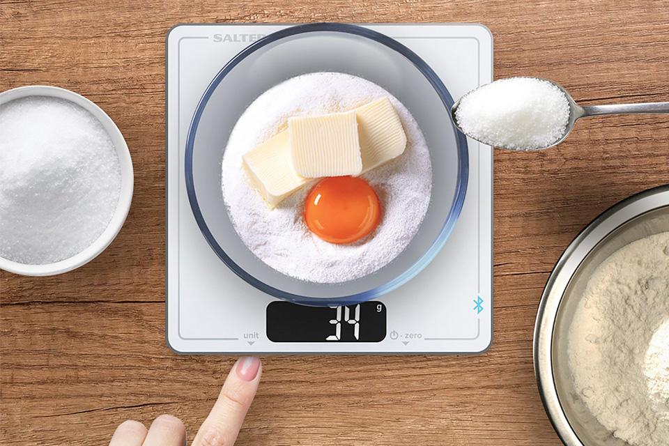 Digital scales weighing sugar, butter and an egg. More sugar is being added by the cook.