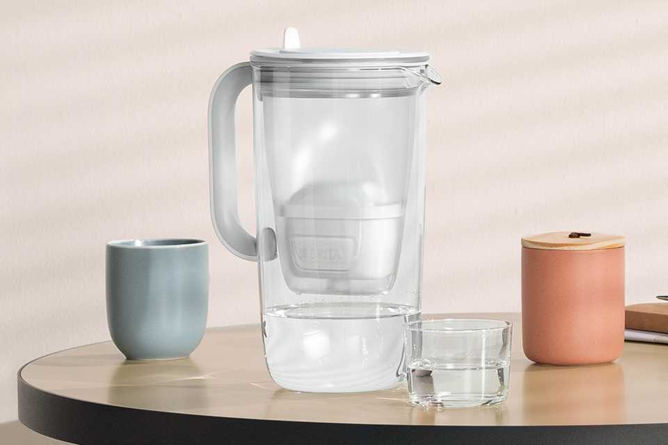 A BRITA glass jug on a table next to a mug and a glass of water.