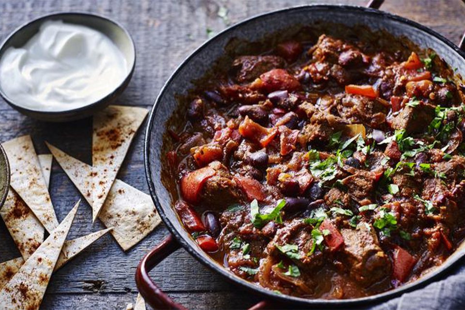 A slow-cooked beef chilli dish served on a table next to a dip and garnish.